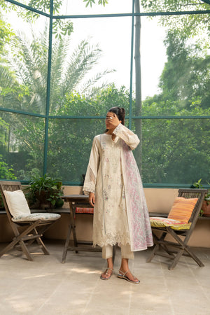 Woman in a light-colored dress standing in a sunlit room with large windows showing greenery outside.