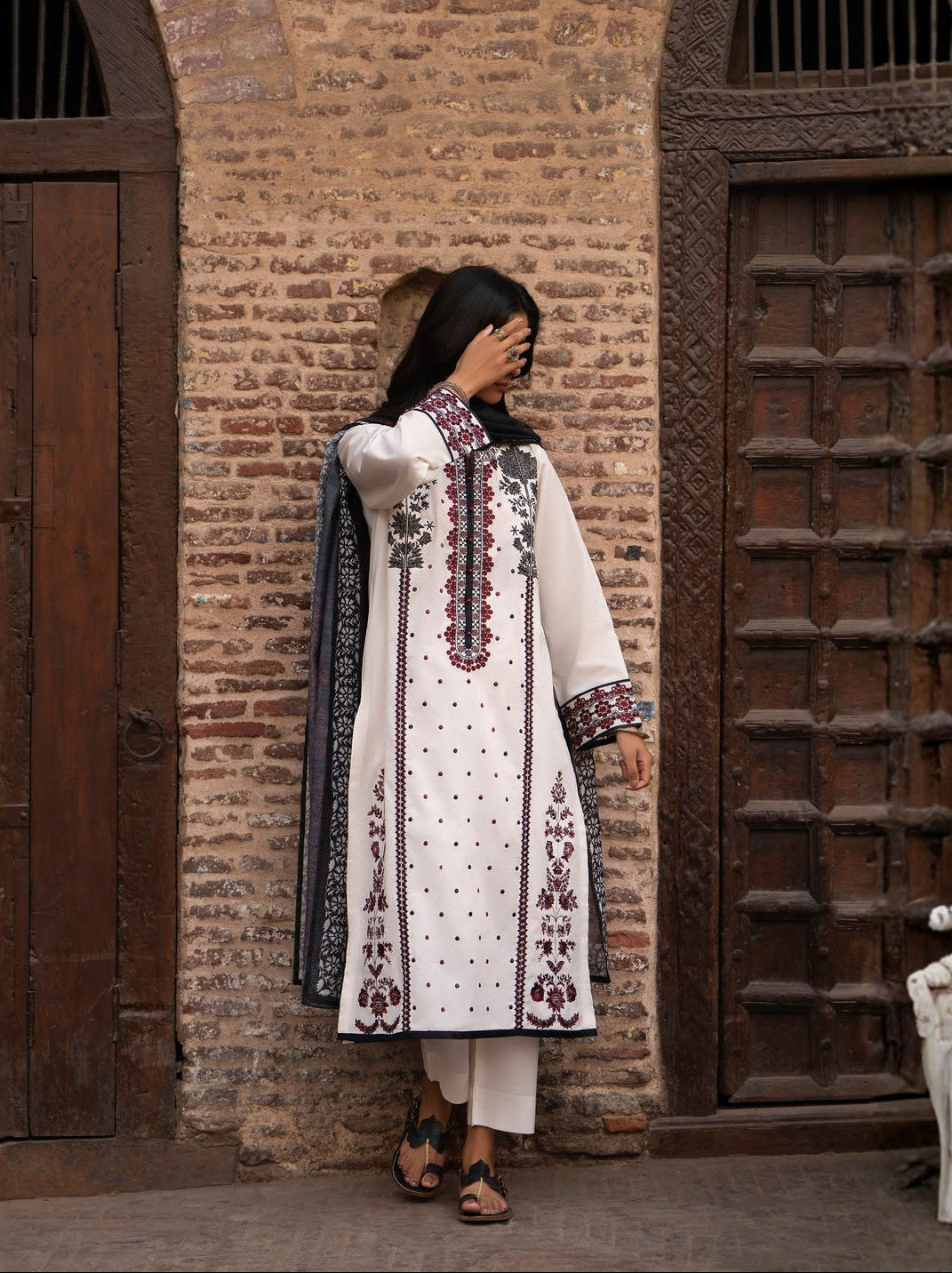 Woman in traditional outfit standing against a brick wall with wooden doors.